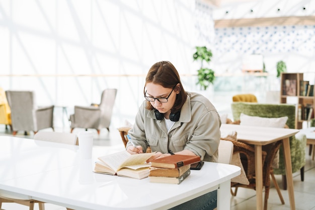 De jonge donkerbruine student van het tienermeisje in glazen die huiswerk met boeken doen bij groene moderne bibliotheek openbare plaats