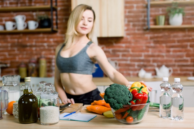 De jonge en gelukkige vrouw is klaar om verse groenten en fruit te eten, zittend op de lijst met gezonde verse ingrediënten in moderne lichte keuken