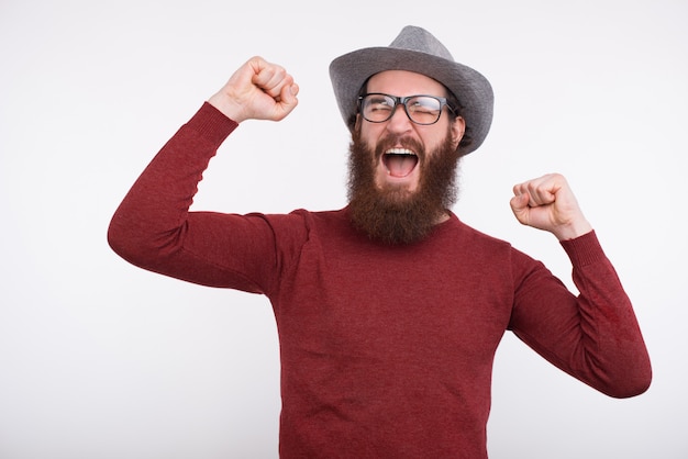 De jonge gebaarde mens met glazen en een grijze hoed schreeuwt met zijn omhoog wapens.