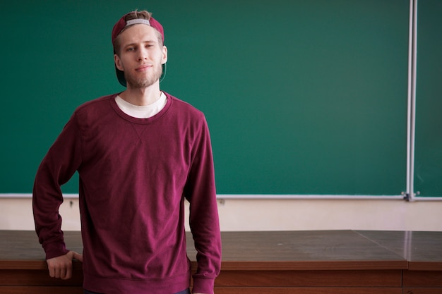 De jonge gebaarde student zit op het bureau van de leraarslijst voor bord met exemplaarruimte