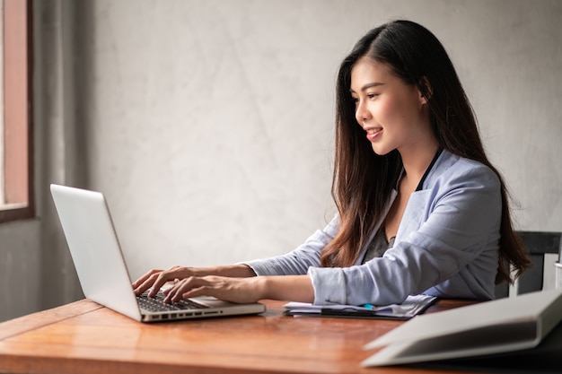 De jonge gelukkige Aziatische onderneemster in blauw overhemd die van huis werken en computerlaptop gebruiken en idee voor haar zaken denken