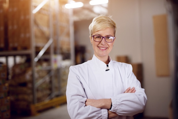 De jonge gelukkige professionele trotse vrouwelijke werknemer in witte steriele doeken houdt haar wapens gekruist en bevindt zich in fabrieksopslagruimte.