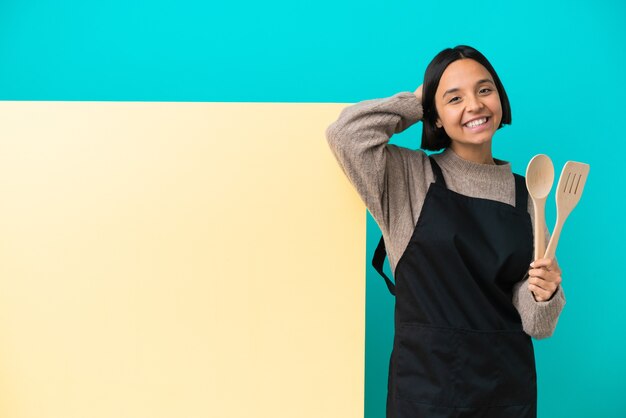 De jonge gemengde vrouw van de raskok met een groot aanplakbiljet dat bij het blauwe muur lachen wordt geïsoleerd