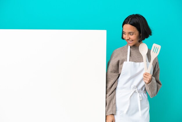 De jonge gemengde vrouw van de raskok met een groot aanplakbiljet dat op blauwe achtergrond wordt geïsoleerd die aan de kant kijkt en glimlacht