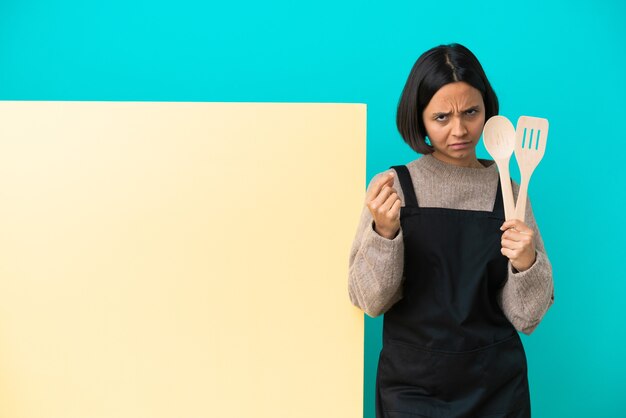 De jonge gemengde vrouw van de raskok met een groot aanplakbiljet dat op blauwe achtergrond wordt geïsoleerd die geldgebaar maakt maar is geruïneerd