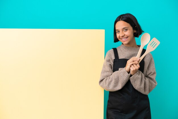 De jonge gemengde vrouw van de raskok met een groot aanplakbiljet dat op blauwe achtergrond wordt geïsoleerd die iets bedenkt