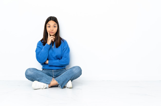 De jonge gemengde zitting van de rasvrouw op de vloer die bij witte muur en het denken wordt geïsoleerd