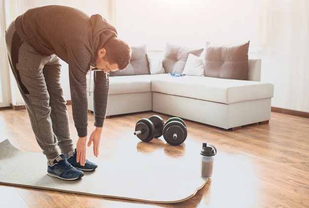 De jonge gewone mens gaat thuis voor sport. Ga op de mat staan en strek je tenen uit. Gewone man die zijn lichaam opwarmt voor het sporten. Alleen trainen in het midden van de kamer. Sportuitrusting op de vloer.