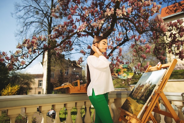 De jonge glimlachende donkerbruine vrouwenkunstenaar schildert een beeld op de straat, dichtbij een mooie boom van magnolia