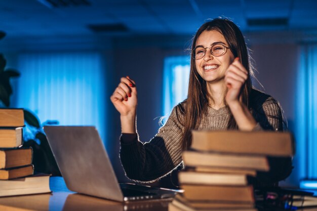 De jonge glimlachende studentenvrouw zit 's avonds aan een tafel in de bibliotheek met een stapel boeken en werkt op een laptop. Voorbereiding op het examen