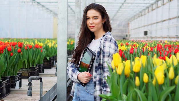 De jonge glimlachende vrouwelijke bloemist bevindt zich met een tablet in haar handen in een kas