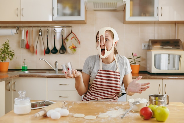 De jonge grappige vrolijke en lachende vrouw legt een deeg met gaten op haar gezicht en heeft plezier in de keuken. Thuis koken. Eten koken.