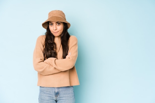 De jonge indische vrouw die een hoed draagt die op blauw wordt geïsoleerd, haalt schouders op en opent verwarde ogen.