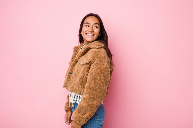 Foto de jonge indische vrouw die een korte schapehuidlaag draagt kijkt opzij glimlachend, vrolijk en aangenaam