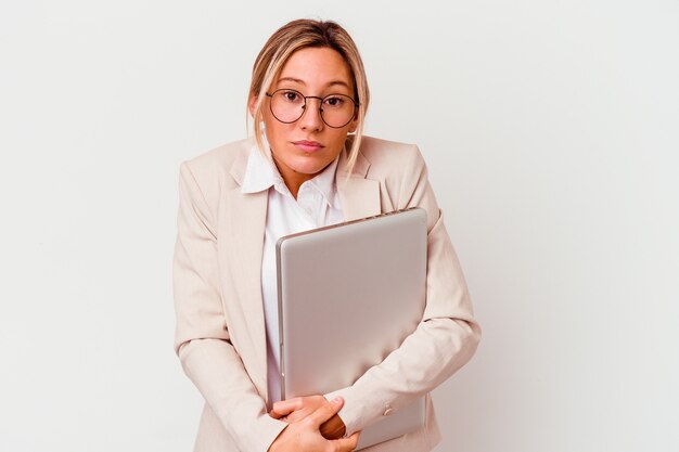 De jonge kaukasische bedrijfsvrouw die laptop houdt die op witte achtergrond wordt geïsoleerd, haalt schouders op en opent verwarde ogen.