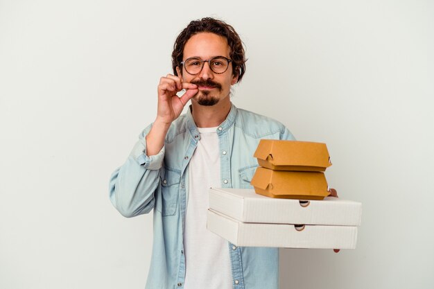 De jonge kaukasische hamburger van de mensenholding pizza's die op wit met vingers op lippen worden geïsoleerd die een geheim houden.