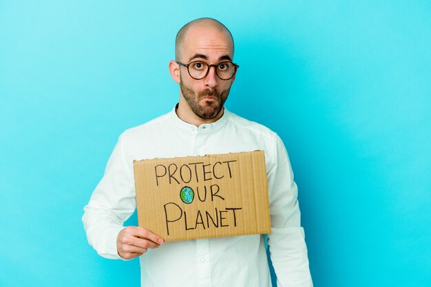 Foto de jonge kaukasische kale mens die een aanplakbiljet beschermt onze planeet houdt dat op purpere muur wordt geïsoleerd, haalt zijn schouders op en opent verwarde ogen.