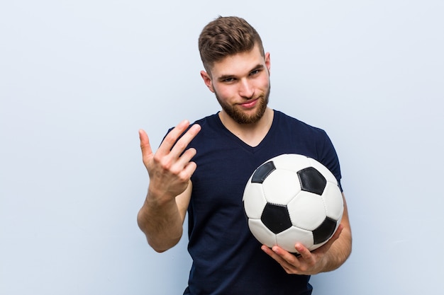 De jonge Kaukasische mens die een voetbalbal houden richtend met vinger op u alsof uitnodigend dichterbij kom.