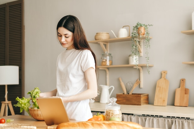 De jonge kaukasische tablet van het vrouwengebruik in de keuken en het koken van salade