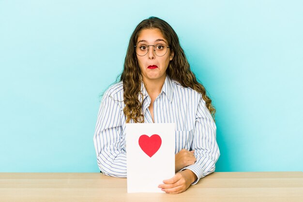 De jonge kaukasische vrouw die een geïsoleerde valentijnskaartkaart houdt haalt schouders op en opent verwarde ogen.