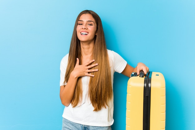 De jonge Kaukasische vrouw die een reiskoffer houdt lacht luid het houden van hand op borst.