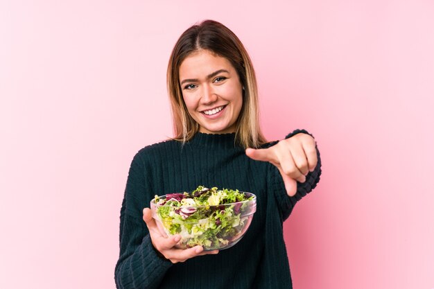 De jonge kaukasische vrouw die een salade houdt isoleerde vrolijke glimlachen die naar voorzijde wijzen.