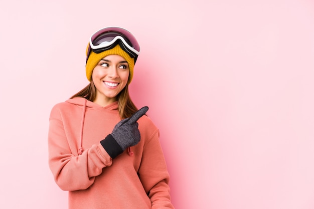 De jonge Kaukasische vrouw die een ski draagt kleedt geïsoleerde glimlachend en opzij richtend, tonend iets op lege ruimte.