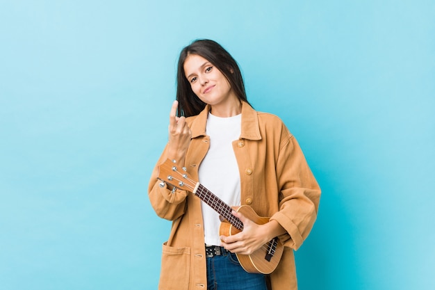 De jonge Kaukasische vrouw die een ukelele houden richtend met vinger op u alsof uitnodigend dichterbij kom.