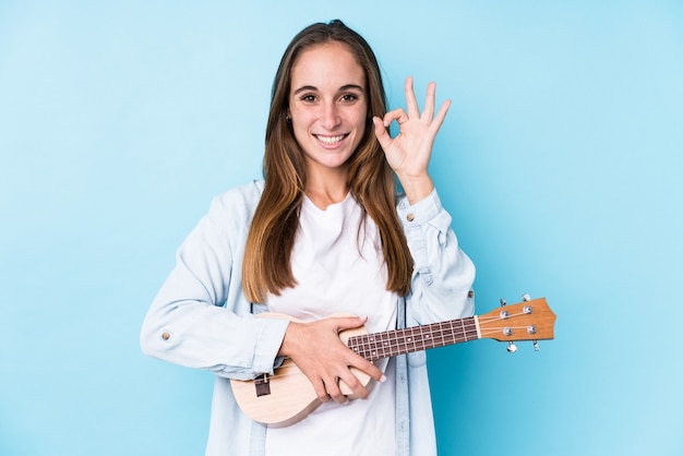 De jonge kaukasische vrouw die een ukelele houdt isoleert vrolijk en zelfverzekerd tonend ok gebaar.