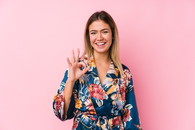 De jonge kaukasische vrouw die pyjama draagt knipoogt een oog en houdt een goed gebaar met hand.