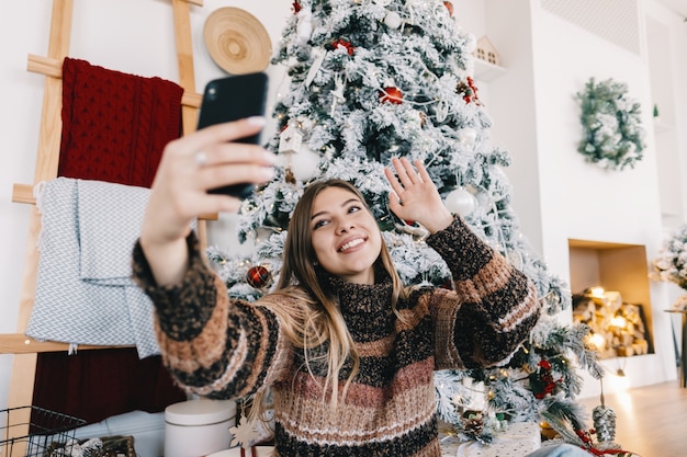 De jonge kaukasische vrouw wenst prettige Kerstmis door videogesprek met behulp van mobiele telefoon in vakantie thuis op de bank.