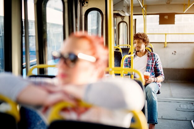 Foto de jonge knappe mens zit op een buszetel terwijl hij koffie houdt en door het raam kijkt terwijl hij op aankomst op zijn bestemming wacht.