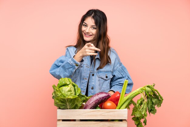 De jonge landbouwersvrouw die een mandhoogtepunt van verse groenten houden richt vinger op u