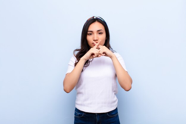 De jonge Latijnse mooie vrouw die ernstig en ontstemd met beide vingers kijken kruiste vooraan in afwijzing, die om stilte tegen vlakke muur vragen