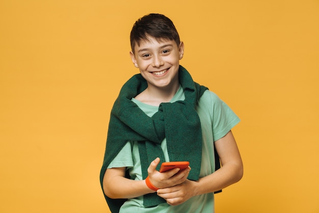 De jonge leuke Kaukasische jongen, brede glanzende glimlach, draagt lichtgroen overhemd, houdt mobiele telefoonmodellen over gele muur. Positief mensenconcept. vakantie begon