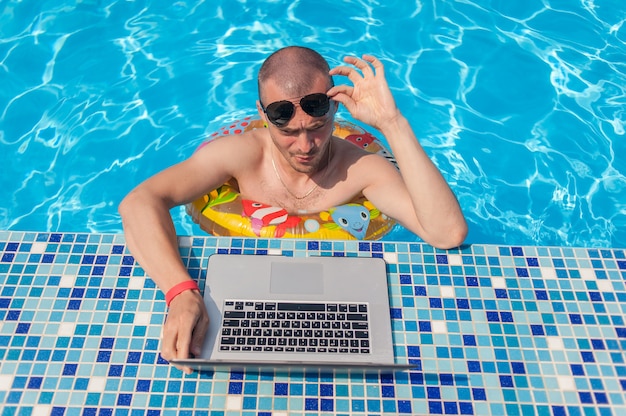 De jonge man op zomervakantie met de laptop, zaken online. afstand thuiskantoor in zwembad in rubberen ring op quarantaine