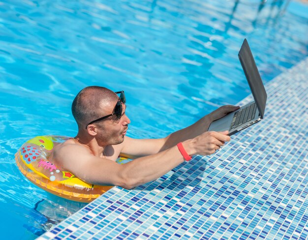 De jonge man op zomervakantie met de laptop, zaken online. afstand thuiskantoor in zwembad in rubberen ring op quarantaine