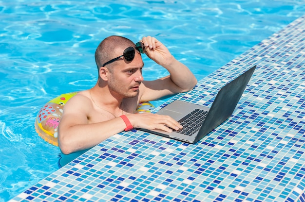 De jonge man op zomervakantie met de laptop, zaken online. Afstand thuiskantoor in zwembad in rubberen ring op quarantaine