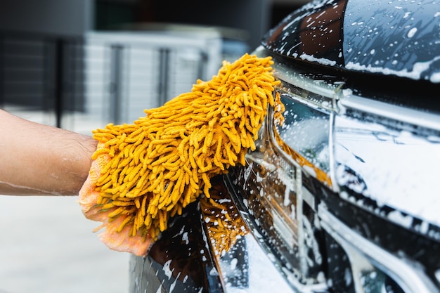 De jonge man waste zijn auto met schuimend reinigingsschuim. Hij droeg handschoenen bij het wassen van de auto. Hij veegde de koplampen van de auto schoon.