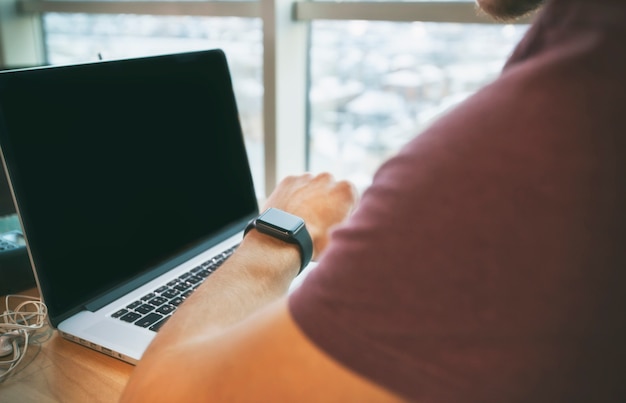 De jonge man zit op de laptop en kijken op de smartwatch