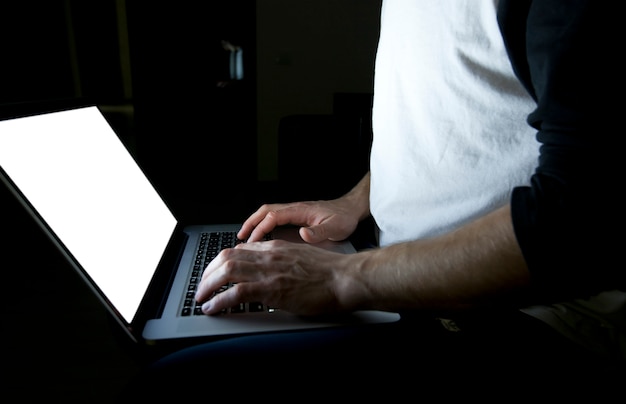 De jonge man zittend op de bank met moderne laptop in de avond en surfen op het web