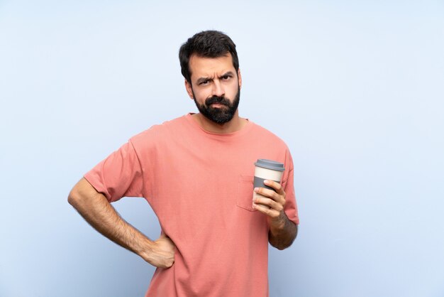 De jonge mens die met baard houden haalt koffie over geïsoleerde blauwe boze muur weg