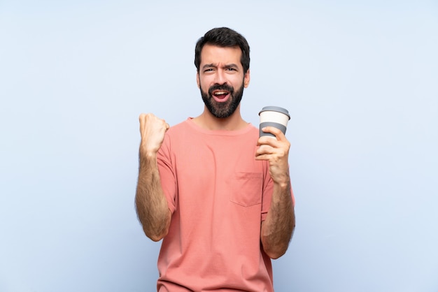 De jonge mens die met baard houdt haalt koffie over geïsoleerd blauw weg het vieren van een overwinning in winnaarpositie