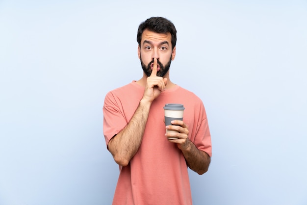 De jonge mens die met baard houdt haalt koffie over geïsoleerde blauwe muur weg die een teken van stiltegebaar tonen die vinger in mond zetten