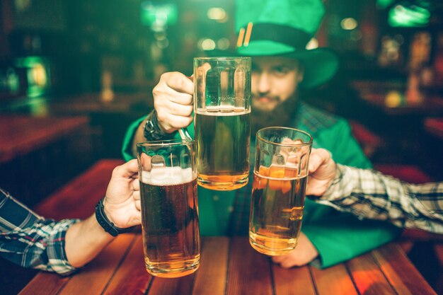 De jonge mens in groen kostuum zit aan tafel met vrienden in bar en houdt biermokken samen. hij is geconcentreerd.