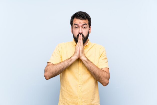 De jonge mens met baard over geïsoleerde blauwe muur houdt palm samen. Persoon vraagt om iets