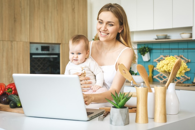 De jonge moeder kookt en speelt met haar babydochter in een moderne keuken het plaatsen. Kijkend naar laptop, glimlachend.