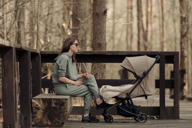 De jonge moeder ontspant op de houten bank terwijl haar kind in de kinderwagen slaapt De moeder met haar kind in het park voor frisse lucht