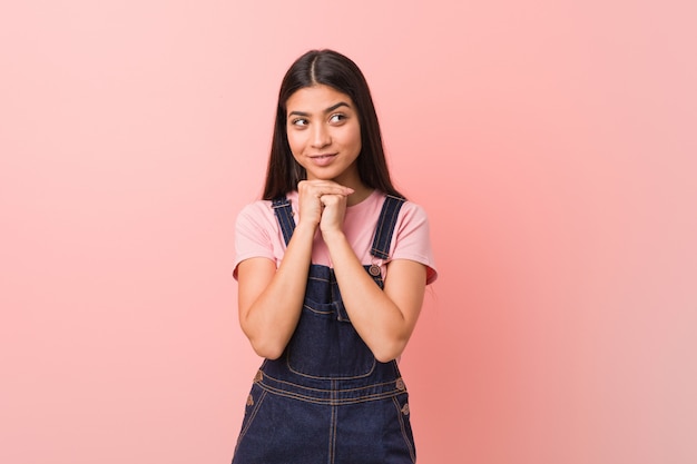 De jonge mooie Arabische vrouw die een jeansdungaree draagt houdt handen onder kin, kijkt gelukkig opzij.