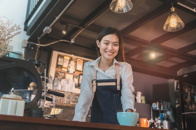 De jonge mooie aziatische barista van de vrouwenbarrière draagt ​​blauwe schort